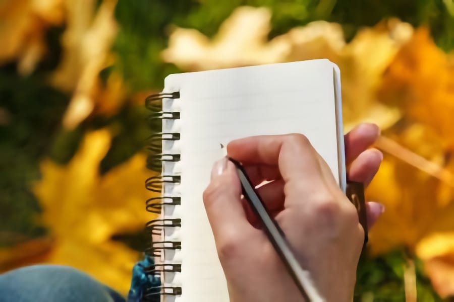 Person writing in a small notebook outdoors with autumn leaves in the background