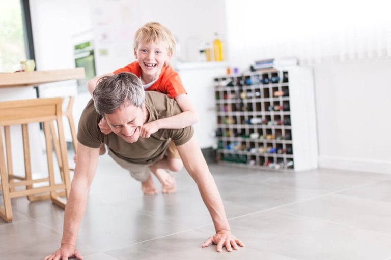 Father doing push-ups with his smiling son on his back in a bright modern home, showing family comfort from reliable HVAC heating and cooling.