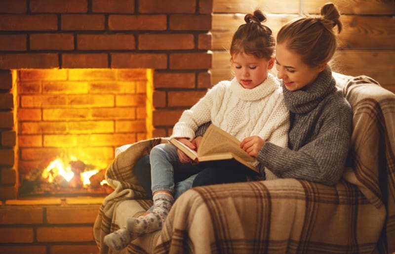 Mother and child sitting together under a blanket by a warm fireplace, reading a book in a cozy living room