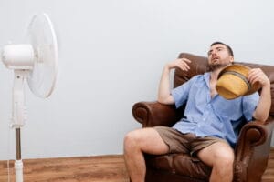 Man sitting in a chair fanning himself and using an electric fan, illustrating discomfort from a broken or inefficient air conditioning system