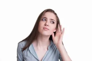 Woman tilting her head and cupping her hand to her ear, listening attentively against a white background
